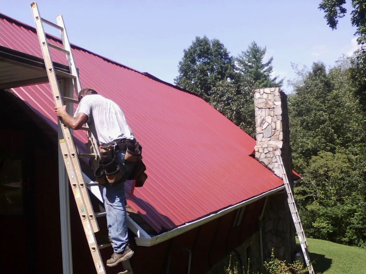 Roofer on a ladder beside a red metal roof and stone chimney — Soffit & Fascia Repair by Roofing Brothers in Winston-Salem